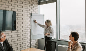 A female Tension Packaging & Automation partner writes on a flip chart during a business meeting while two customers sit at a conference table, listening. Large windows and a brick wall are in the background.